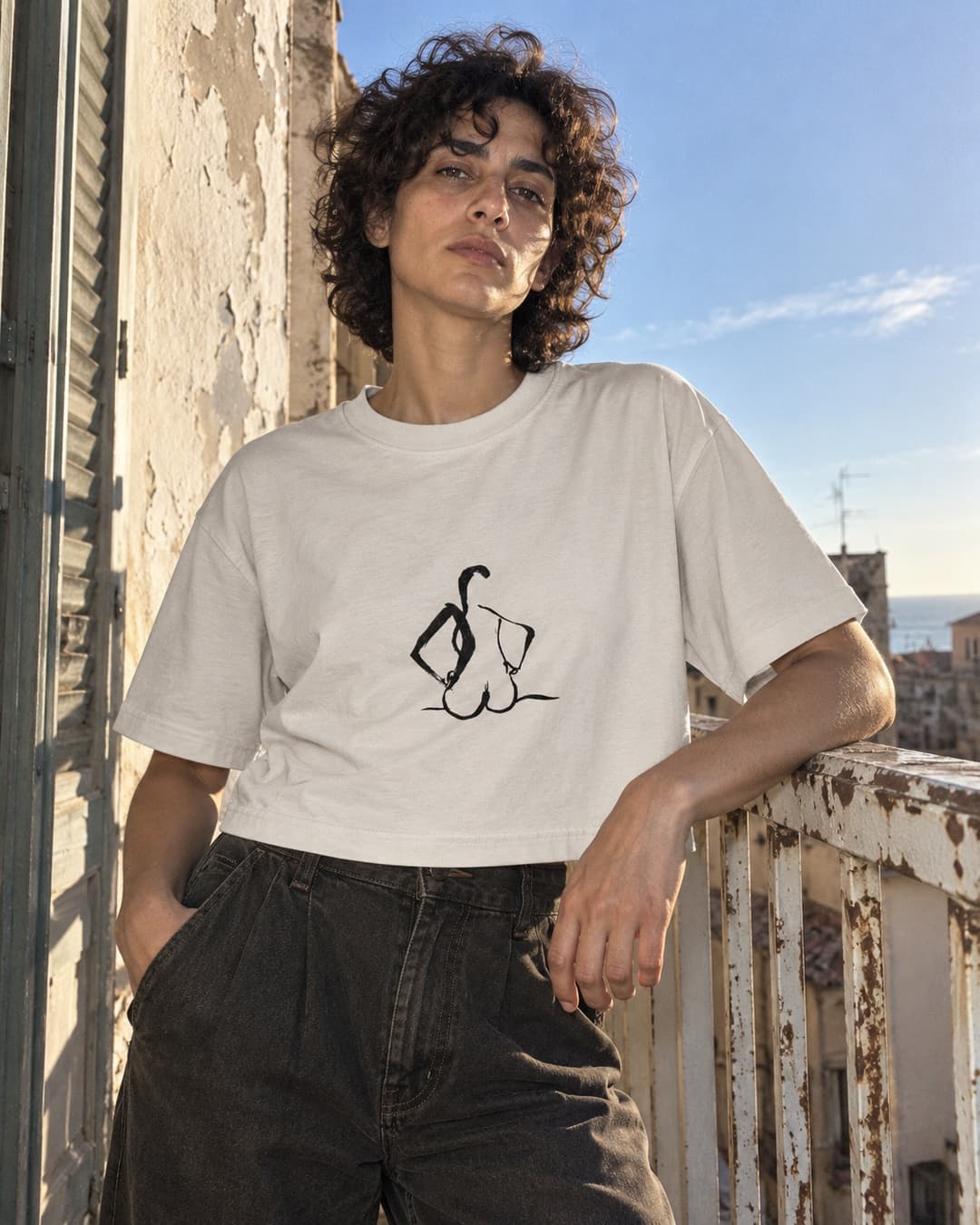 Faded Bone Hands on Hips cropped tee with black ink drawing, worn with dark trousers on a weathered sunny balcony