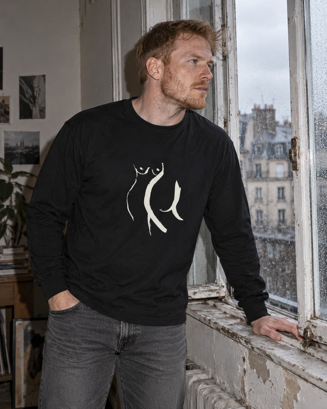 Black Front and Back long-sleeve tee with cream drawing, worn by a man leaning into an open rainy window