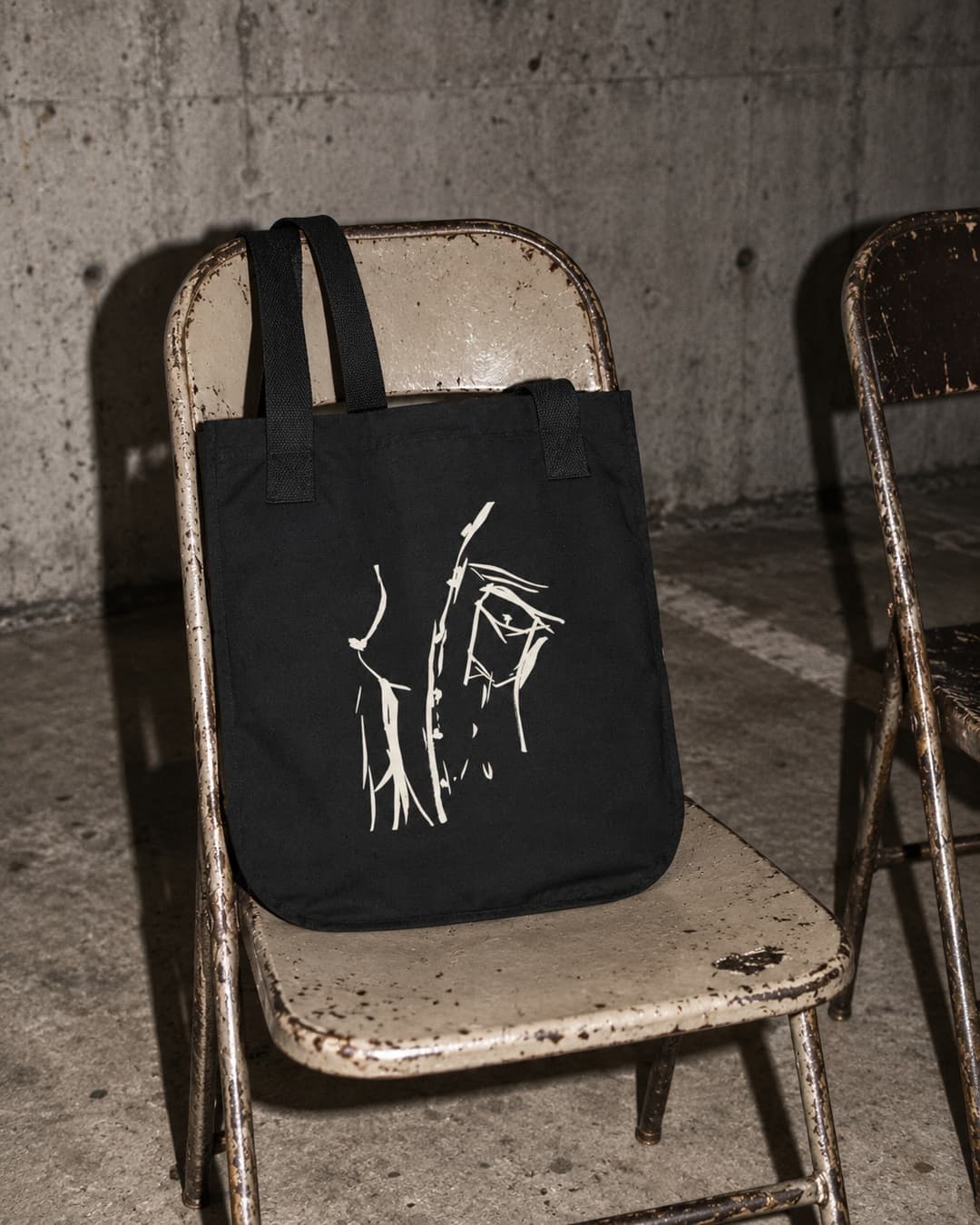 Black Peaking Out canvas tote with cream drawing sitting on a battered folding chair beside an empty chair in a concrete carpark